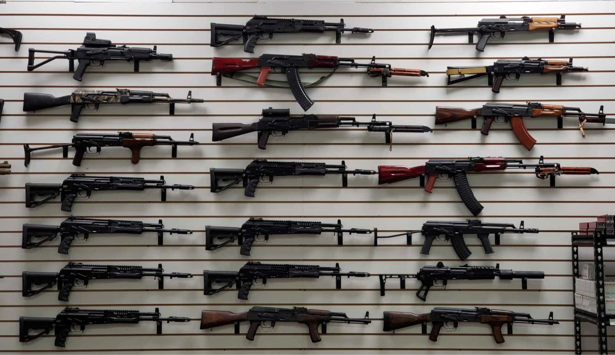 A wall display featuring multiple rifles with wooden and black finishes, arranged in rows on horizontal slats. The rifles vary slightly in size and style but share a similar design.