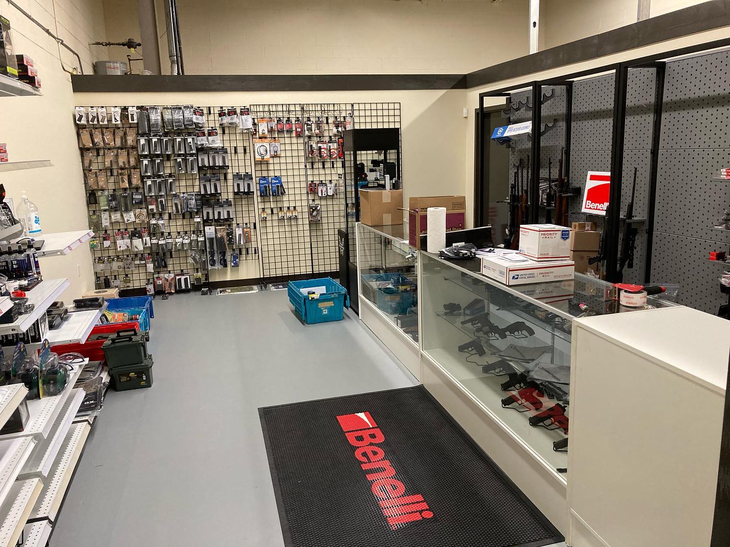 A gun shop interior with firearms and accessories displayed on shelves and racks, glass counters with handguns and boxes, and a black floor mat with a red Benelli logo near the entrance.