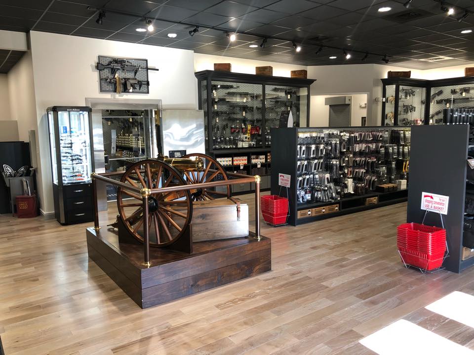 A modern gun store interior with wooden floors, display cases of firearms and accessories, and a large antique cannon on a wooden platform in the center. Red shopping baskets are stacked near the entrance.