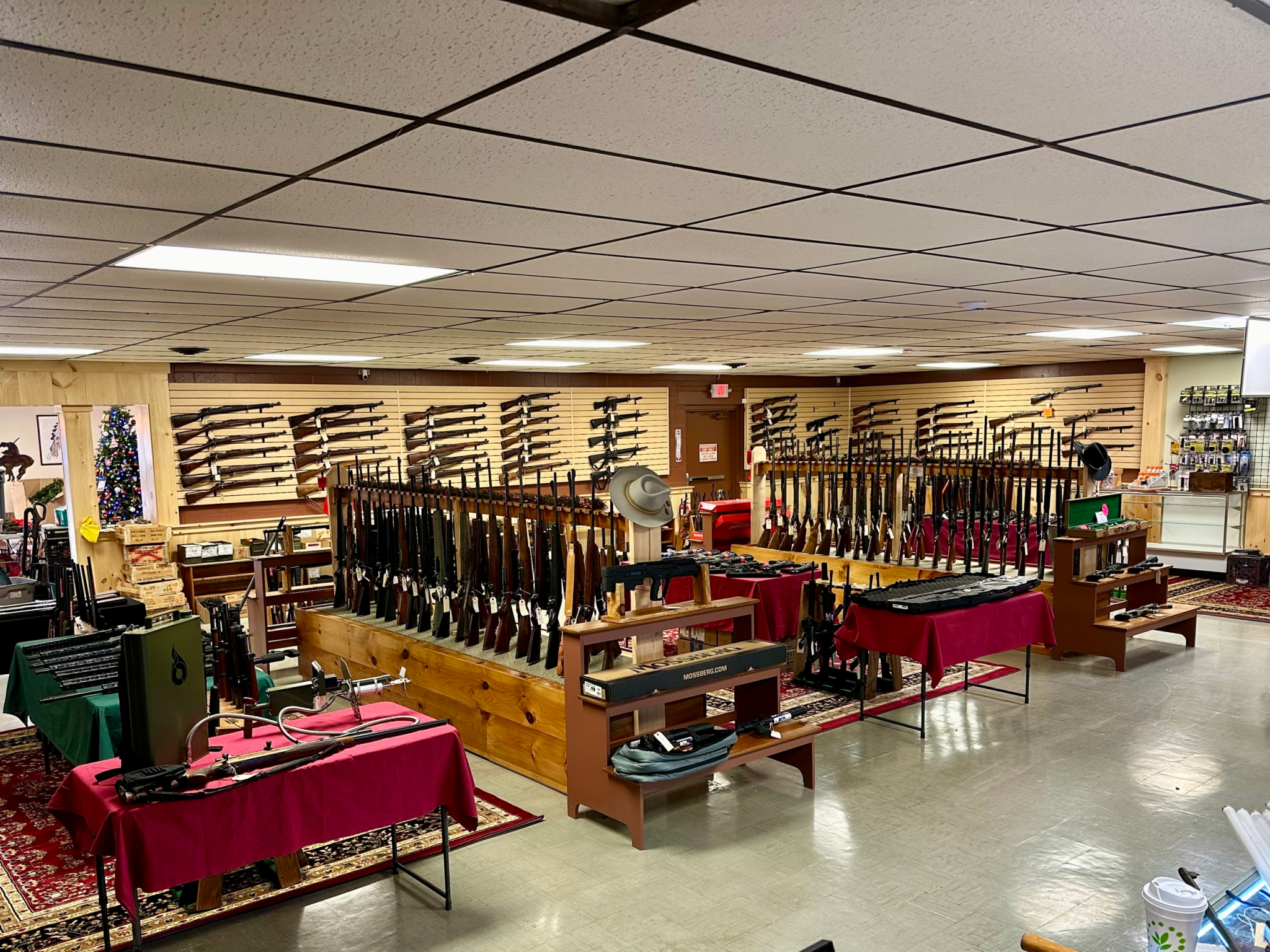 A gun shop interior with rifles and shotguns displayed on wall racks and wooden stands, tables covered with red cloths, and various accessories and items on shelves around the room.