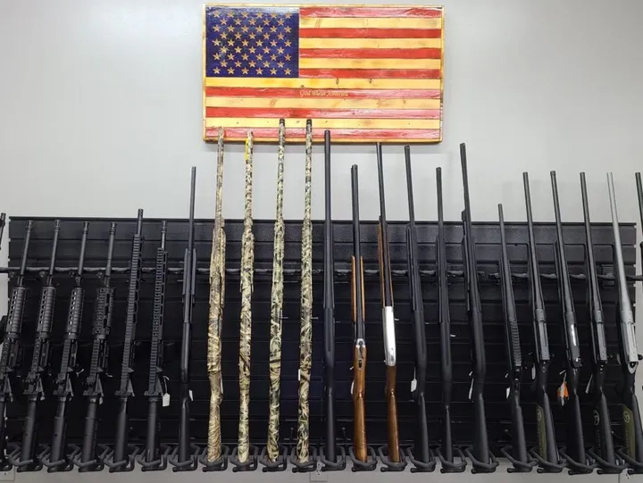 A display of various rifles and shotguns is arranged upright on a rack beneath a wooden American flag on a white wall.