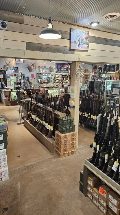 Interior of a firearm store with rifles displayed on stands and shelves. Boxes of ammunition are stacked nearby. The store has wooden walls, a corrugated metal ceiling, and various hunting accessories on display. Hanging lights illuminate the space.