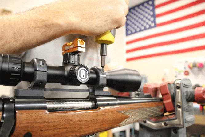 Person working on a rifle scope alignment with tools, inside a workshop. An American flag is visible in the background.