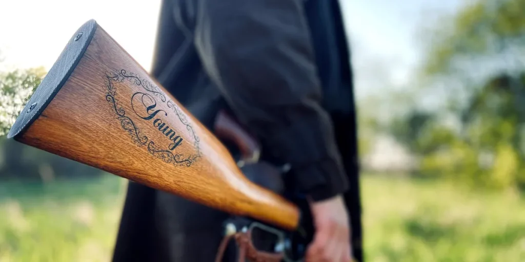 A person stands outdoors holding a rifle by their side. The wooden stock of the rifle is engraved with the name "Young" and decorative scrollwork. The background is blurred greenery and sunlight.