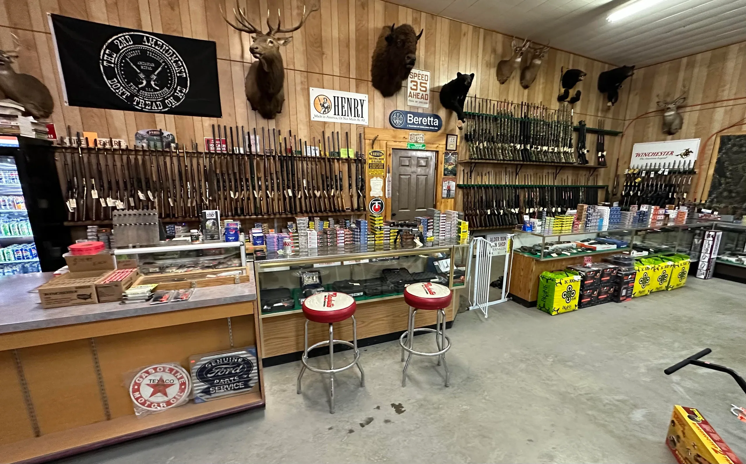 A gun shop interior with rifles displayed on wooden walls, ammunition boxes on glass counters, mounted deer heads, black stools, vintage signs, and snacks and drinks along the side.