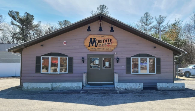 A single-story building with a brown exterior and a sign reading "Mustard Gun Company Indoor Shooting Range" above the entrance, situated in a parking lot with trees in the background.