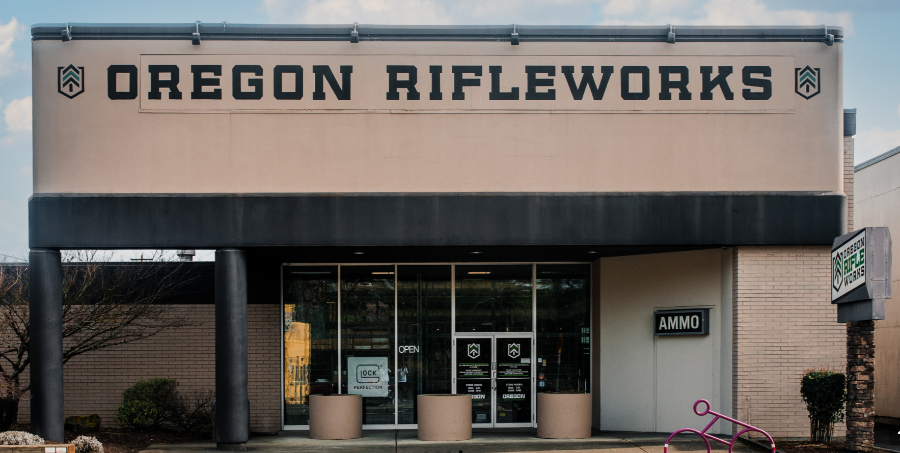 A storefront with a large sign reading "Oregon Rifleworks" above glass doors. The entrance has "AMMO" and "OPEN" signs, and a pink bicycle rack is visible near the entrance.