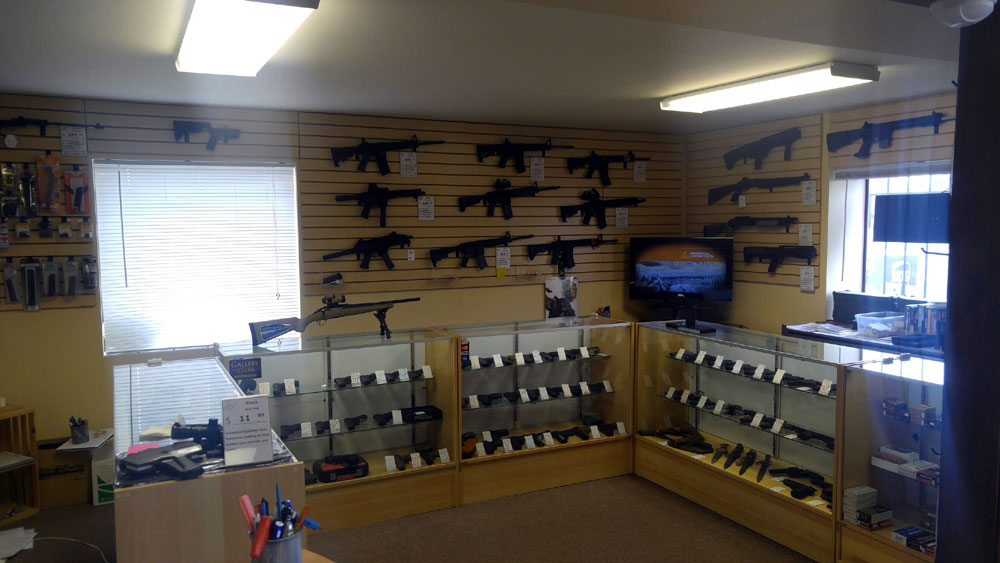 A gun store interior with rifles mounted on the walls, handguns displayed in glass cases, and accessories on shelves. Fluorescent lights illuminate the room and a window lets in natural light.