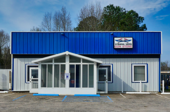A small, blue-and-white building with a sign reading "Strong Arms Gun Store" above the entrance, flanked by trees and a clear sky, with two accessible parking spaces in front.