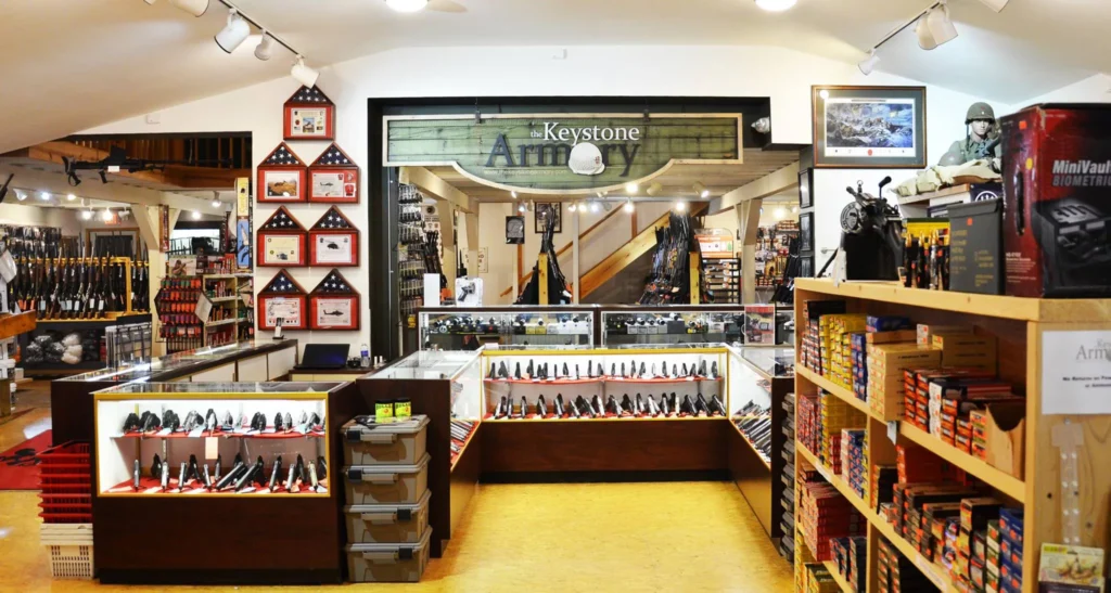 Interior of a gun store with glass display cases containing handguns, shelves stocked with ammunition, rifles on racks, American flags on the wall, and a sign reading "Keystone Armory" above a doorway.