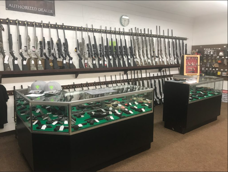 A gun shop interior displaying rifles on wall racks, handguns in glass display cases, and firearm accessories on wall shelves. Two hats are also displayed on one of the cases. A clock is mounted above the rifles.