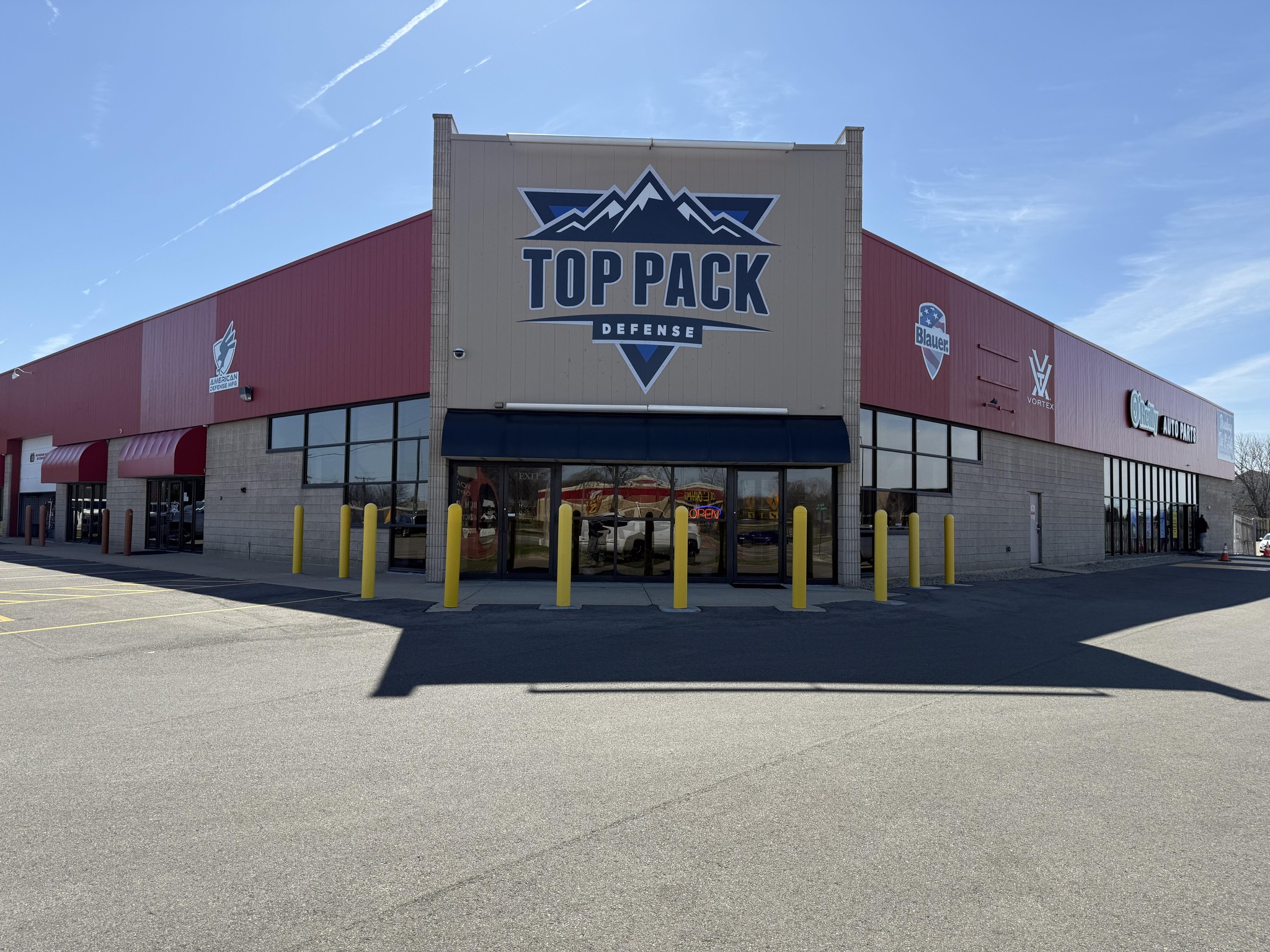 A large commercial building with a sign reading "Top Pack Defense" on the front facade. The building has red accents, glass doors, and yellow bollards at the entrance. The sky is clear and blue.