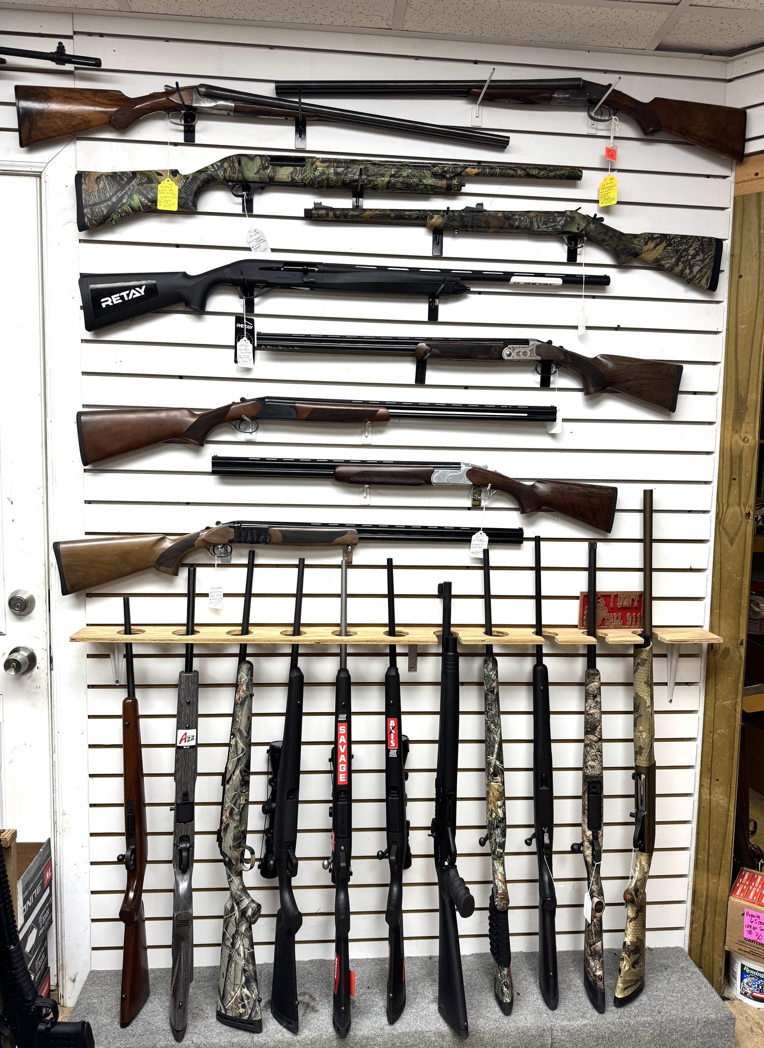 Several rifles and shotguns are displayed on a white slat wall, with more firearms standing upright on a rack below. Some firearms have camouflage patterns; price tags are attached to a few. Boxes of ammo are visible on the shelf.