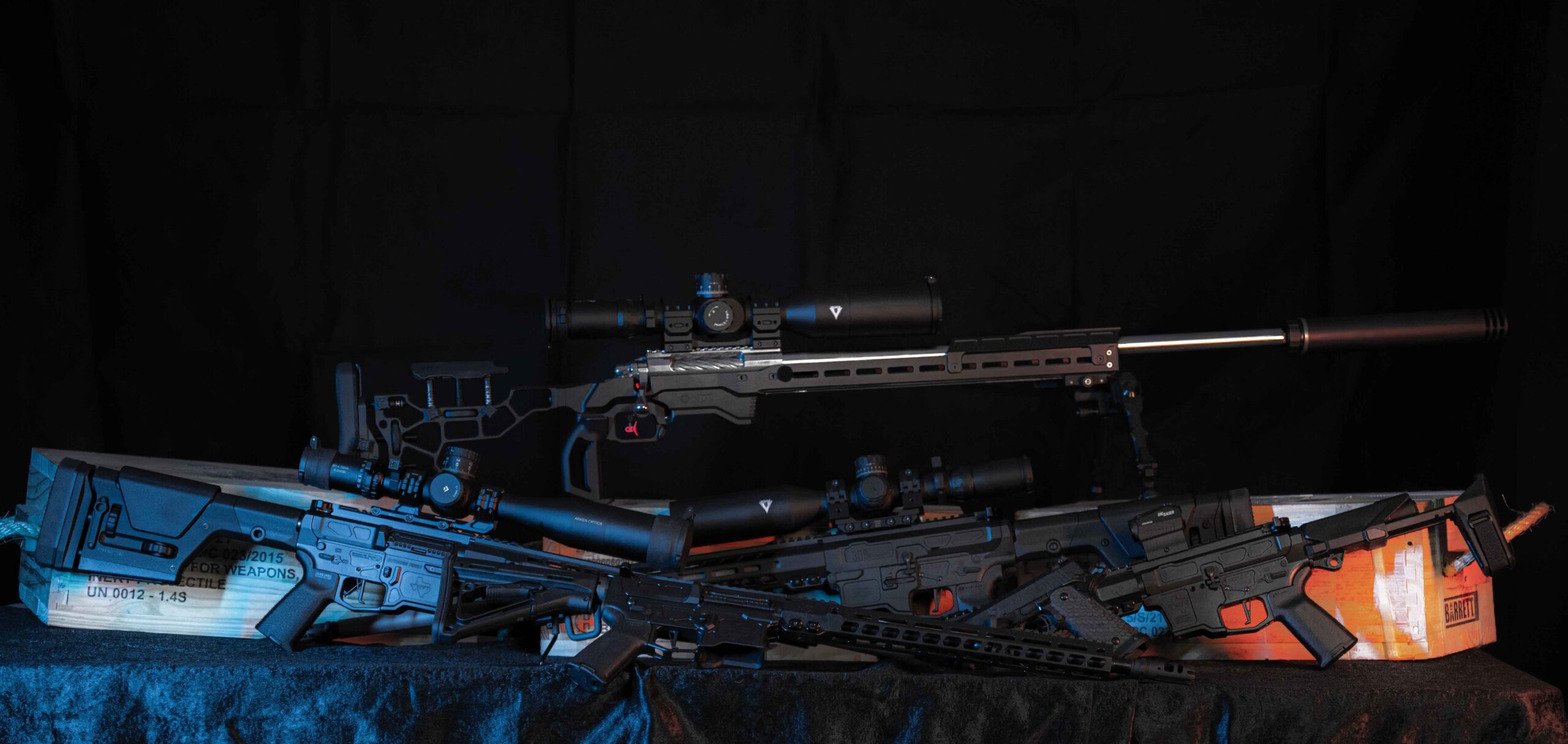 Three modern rifles with scopes and tactical accessories are displayed on a black surface, with a wooden crate beneath them, all set against a dark background with blue and orange lighting accents.