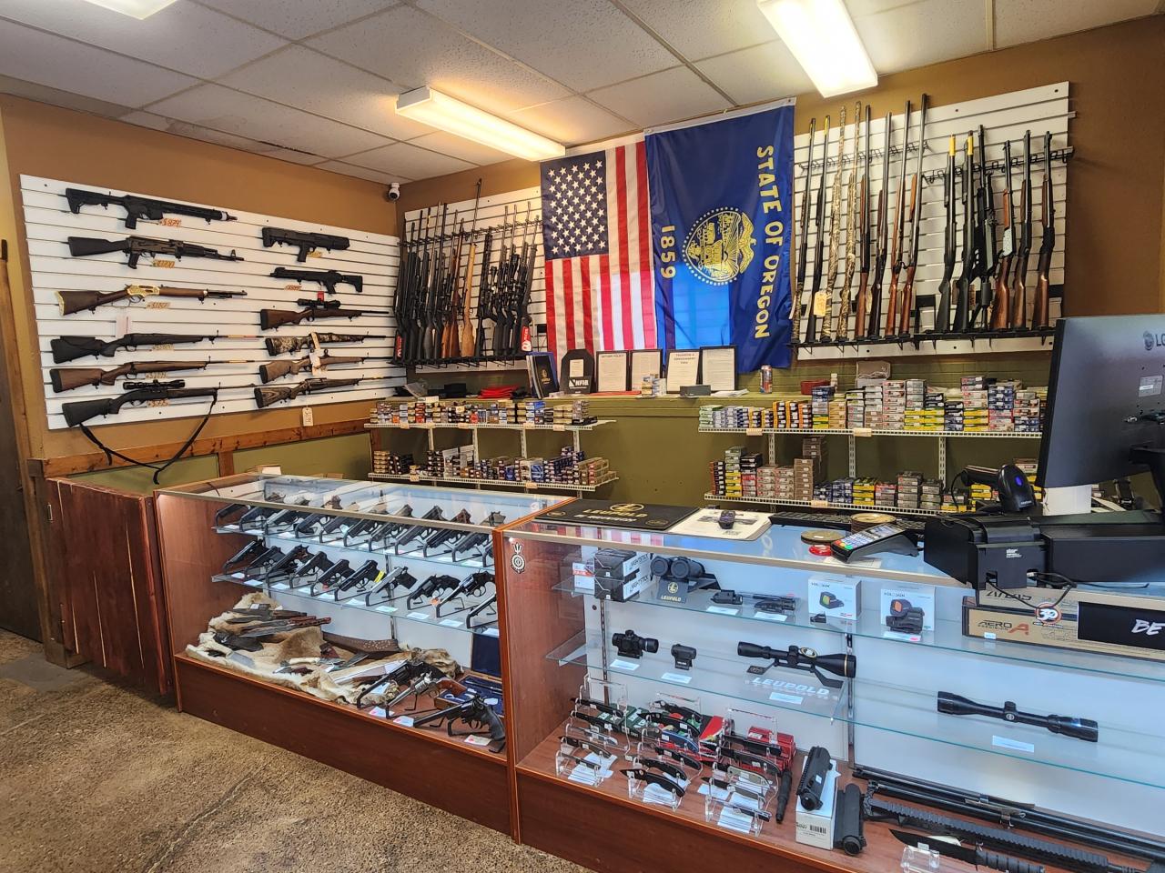 A gun shop interior with rifles and shotguns displayed on the wall, handguns in glass cases, various ammunition boxes on shelves, and an American flag and Oregon state flag hanging behind the counter.
