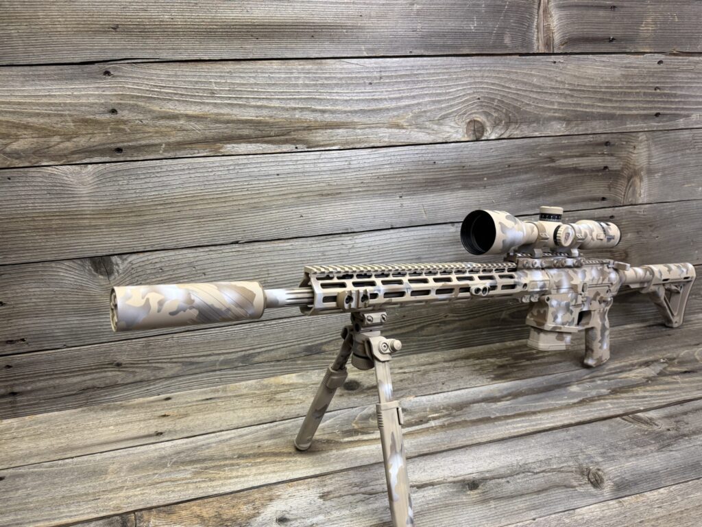 A camouflaged rifle with a scope and bipod, featuring a tan and gray pattern, is positioned on a wooden surface with a matching wood-plank background.