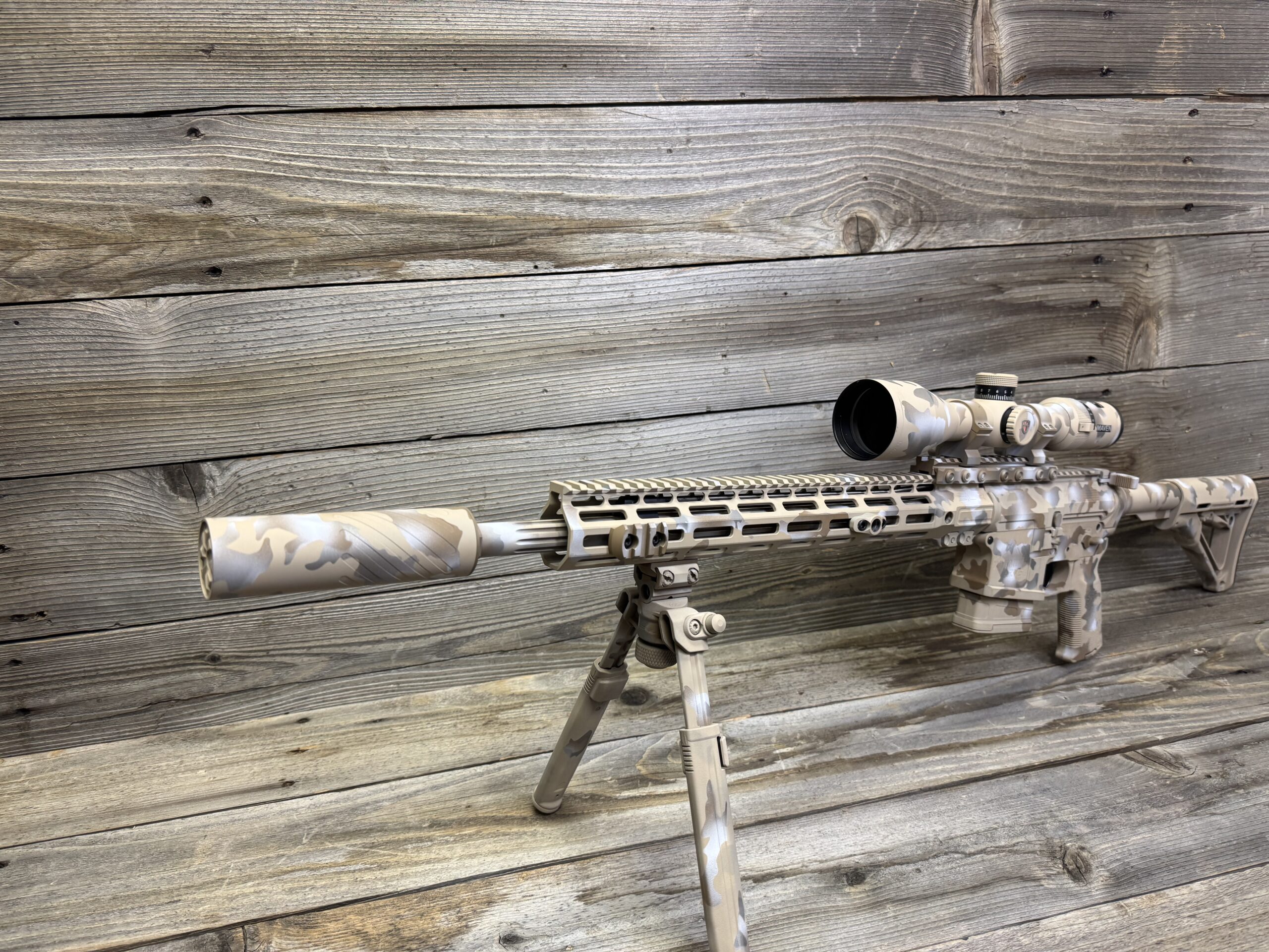 A camouflaged rifle with a scope and bipod, featuring a tan and gray pattern, is positioned on a wooden surface with a matching wood-plank background.