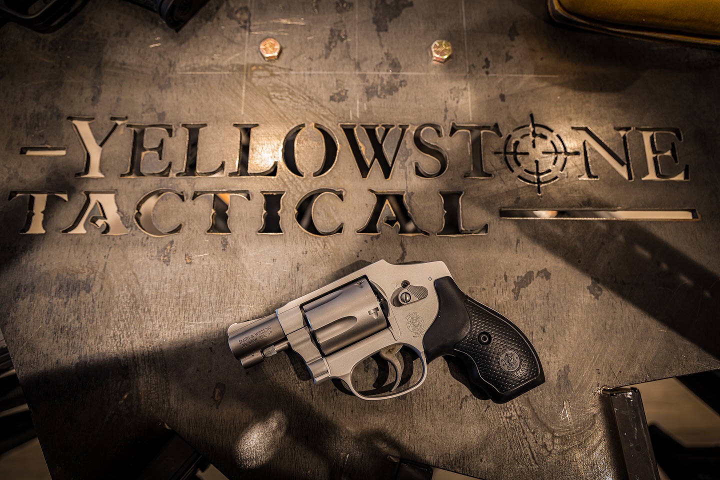 A silver and black revolver rests on a metal surface cut with the words "Yellowstone Tactical" and a target symbol. Two bolts are also visible on the table.
